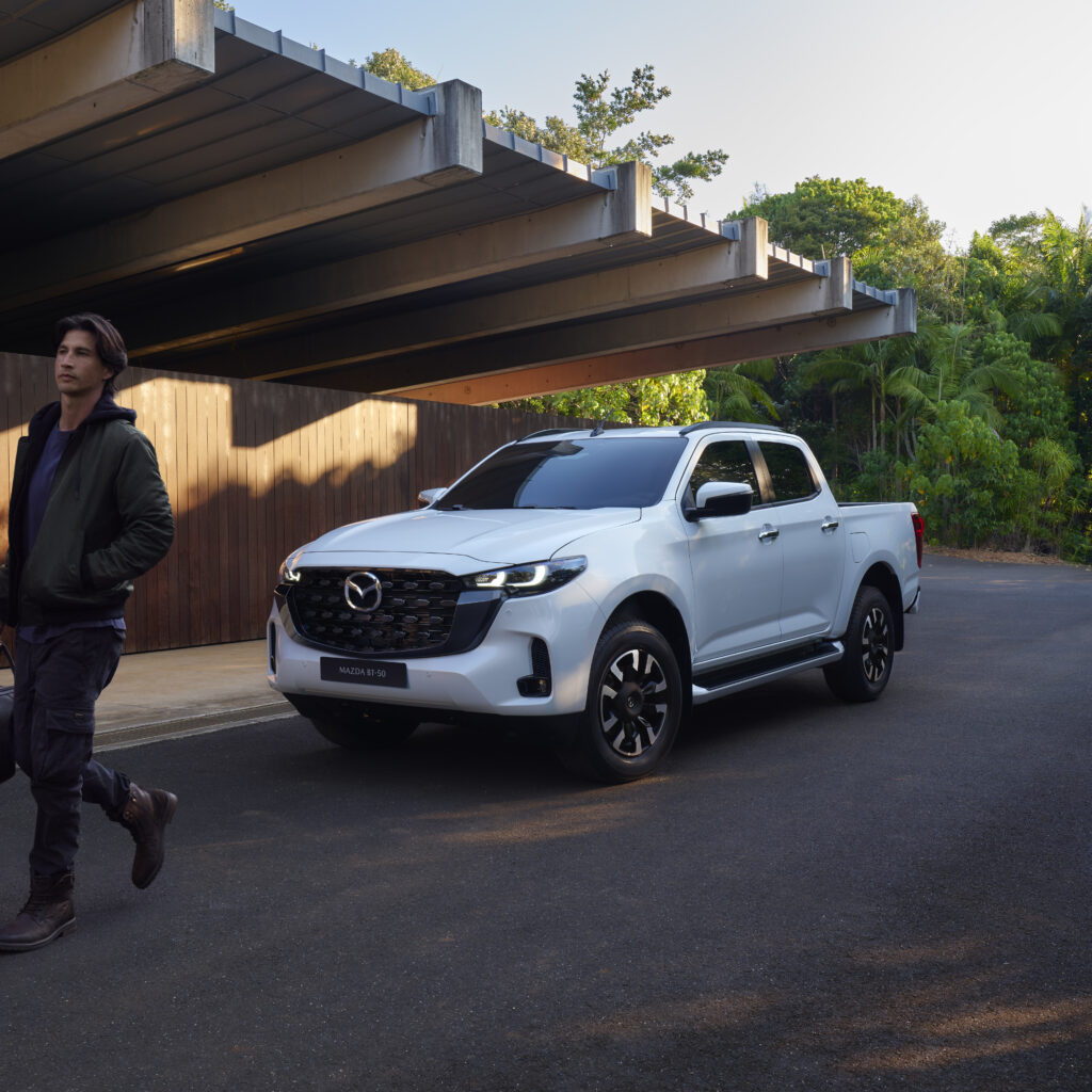 Mazda BT-50 2025 blanca estacionada frente a una casa moderna, junto a un hombre caminando con una maleta.